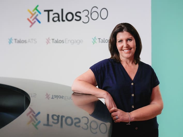 Talos360 Chief Executive Officer, Janette Martin standing in Talos360's office with a wall with Talos360's logo in the background.